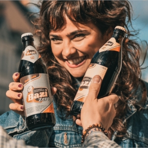 Eine lachende Frau mit lockigem braunem Haar hält eine Flasche „Bazi Zero“ in der Hand und stößt damit fröhlich mit einer weiteren Person an, die ebenfalls eine Flasche „Bazi“ hält. Beide tragen lässige Kleidung und genießen sichtlich einen sonnigen Moment im Freien. Die Stimmung ist ausgelassen und freundlich.