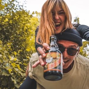 Ein junger Mann trägt eine Frau auf dem Rücken. Beide lachen ausgelassen in die Kamera. Der Mann hält eine Flasche Lösch-Zwerg in der Hand, die in den Vordergrund gestreckt wird. Im Hintergrund sind grüne Bäume zu sehen.