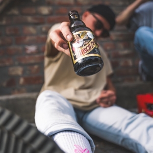Ein junger Mann sitzt lässig vor einer Backsteinwand, streckt den Arm nach vorn und hält eine Lösch-Zwerg-Flasche direkt in die Kamera. Im Vordergrund ist sein Sneaker und weißer Socken mit pinkem Motiv zu sehen.
