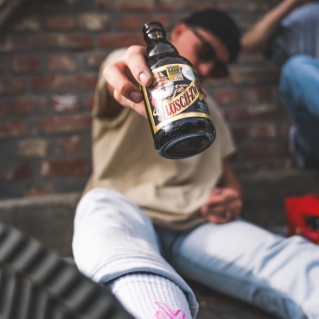 Ein junger Mann sitzt lässig vor einer Backsteinwand, streckt den Arm nach vorn und hält eine Lösch-Zwerg-Flasche direkt in die Kamera. Im Vordergrund ist sein Sneaker und weißer Socken mit pinkem Motiv zu sehen.