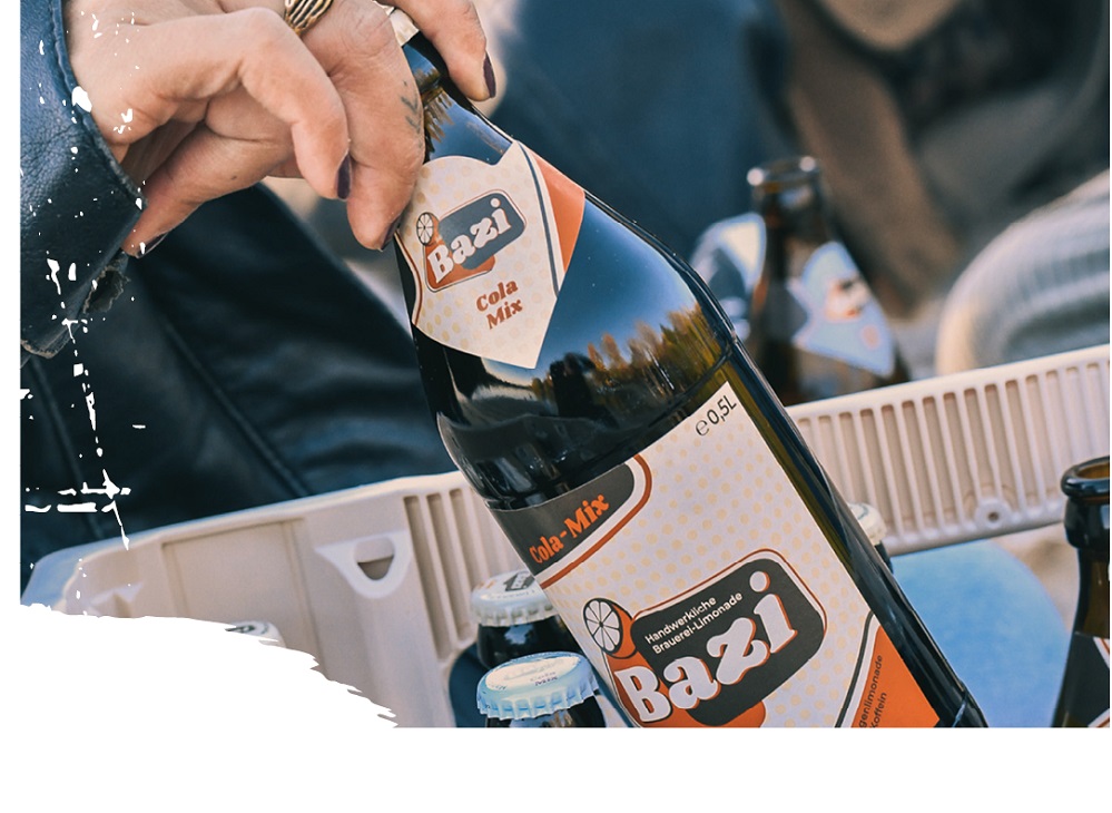 ine Hand greift eine 0,5-Liter-Glasflasche Bazi Cola-Mix aus einer Getränkekiste. Die Flasche hat ein auffälliges Etikett in orange-beige mit der Aufschrift „Bazi – Handwerkliche Brauerei-Limonade“. Im Hintergrund sind weitere Flaschen in der Kiste zu erkennen. Links und unten am Bildrand sind weiße grafische Pinselstriche als Gestaltungselemente eingefügt.
