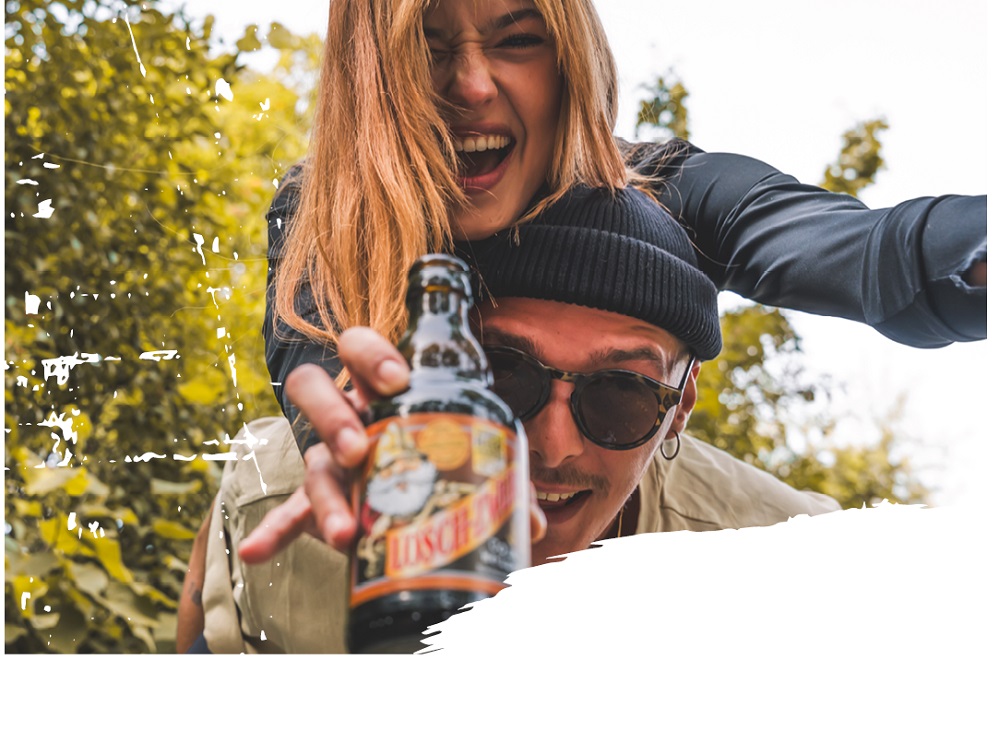 Ein junger Mann trägt eine Frau auf dem Rücken. Beide lachen ausgelassen in die Kamera. Der Mann hält eine Flasche Lösch-Zwerg in der Hand, die in den Vordergrund gestreckt wird. Im Hintergrund sind grüne Bäume zu sehen.