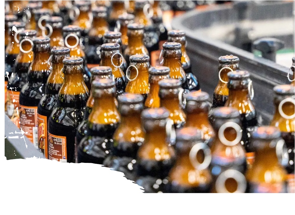 Mehrere Reihen brauner „Lösch-Zwerg“-Bierflaschen mit typischem Metallringverschluss auf einem Förderband in einer Abfüllanlage. Die Flaschen sind ordentlich aufgereiht, die Vorderseiten sind nicht zu sehen – stattdessen Blick auf die Rücketiketten mit Inhaltsangaben. Die Szene vermittelt den industriellen Abfüllprozess in einer Brauerei. Weiße Spritzer am Bildrand deuten auf ein gestalterisches Element im Design hin.