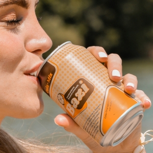 Eine junge Frau mit weiß lackierten Fingernägeln trinkt aus einer orange-beige gepunkteten Dose der Marke „Bazi“. Die Aufschrift „Cola-Mix“ und das Logo mit einer stilisierten Zitronenscheibe sind deutlich sichtbar. Im Hintergrund ist unscharf eine grüne Naturkulisse mit Bäumen und Wasser zu erkennen.
