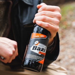 Eine Person in beiger Hose und schwarzer Lederjacke hält eine Flasche „Bazi Energy“ Cola-Mix in der Hand. Die Flasche ist schwarz mit einem auffälligen Etikett in Orange, Weiß und Grau gestaltet. Im Hintergrund ist ein unscharfer Naturweg zu erkennen, was auf eine Outdoor-Szene hinweist.