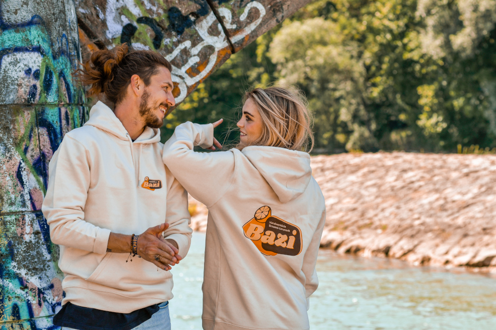 Ein junger Mann und eine junge Frau stehen entspannt an einem bunt besprühten Betonsockel in der Natur am Wasser. Beide tragen helle Hoodies mit dem Logo der Marke „Bazi“ – der Mann auf der Brust, die Frau auf dem Rücken. Sie lachen sich freundlich an und genießen den sonnigen Moment. Die Szene vermittelt Leichtigkeit, Freundschaft und einen natürlichen Lifestyle.