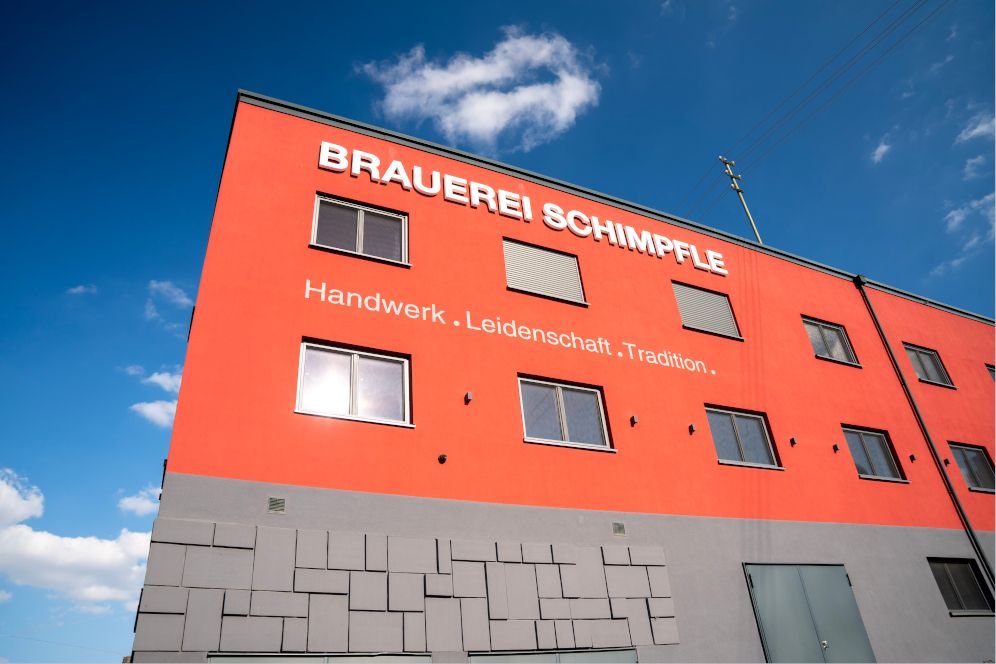 Das Bild zeigt die Außenfassade eines modernen Gebäudes in kräftigem Rot mit grauem Sockelbereich. Auf der Fassade ist in großen weißen Buchstaben der Schriftzug „BRAUEREI SCHIMPFLE“ zu sehen. Darunter steht der Slogan: „Handwerk . Leidenschaft . Tradition .“ Die Kombination aus Farbe, klarer Typografie und dem traditionellen Slogan vermittelt den Eindruck eines regional verwurzelten, handwerklich arbeitenden Unternehmens mit langer Brautradition – eine familiengeführte Brauerei. Das strahlend blaue Himmelsbild unterstreicht die einladende und professionelle Atmosphäre.