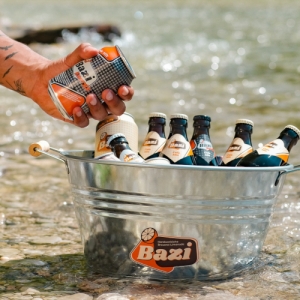 Eine Hand nimmt eine Bazi Energy Cola-Mix-Dose aus einem metallenen Getränkekübel mit mehreren Bazi-Flaschen, der im flachen Wasser eines Flusses steht.