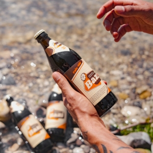 Eine tätowierte Hand hält eine Bazi Cola-Mix-Flasche über einem flachen, steinigen Gewässer. Im Hintergrund liegen mehrere weitere Flaschen halb im Wasser.