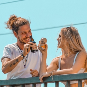 Ein junger Mann und eine junge Frau stoßen lachend mit einer Flasche und einer Dose Bazi an. Sie lehnen an einem Geländer unter blauem Himmel und genießen gemeinsam den Moment.