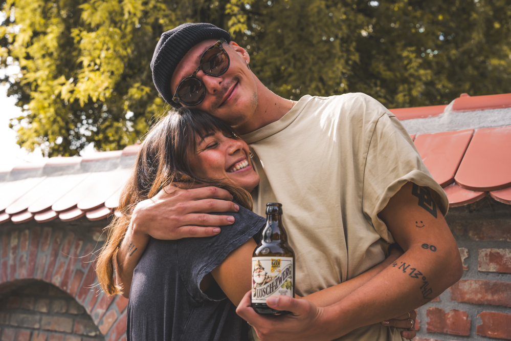 Ein junger Mann mit Sonnenbrille und Mütze umarmt herzlich eine lachende junge Frau. Beide stehen vor einer Mauer mit roten Dachziegeln. Der Mann hält eine Lösch-Zwerg-Flasche in der Hand.