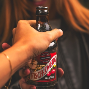Eine Hand reicht einer lachenden jungen Frau eine Flasche Lösch-Zwerg Goiß. Der Fokus liegt auf der Flasche mit dem roten Etikett, der Hintergrund ist unscharf.
