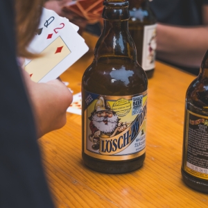 Eine Person spielt Karten an einem Holztisch, auf dem mehrere Flaschen Lösch-Zwerg stehen. Der Fokus liegt auf der Flasche Lösch-Zwerg Weizen Zitrone.