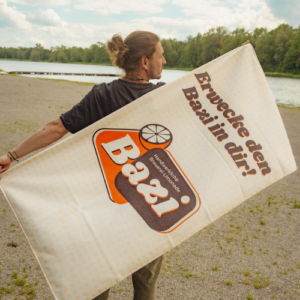 Person steht am Seeufer und hält ein großes, beigefarbenes Handtuch mit dem Bazi-Logo und dem Schriftzug "Erwecke den Bazi in dir!". Im Hintergrund sind Kiesstrand, Wasser und Bäume zu sehen.
