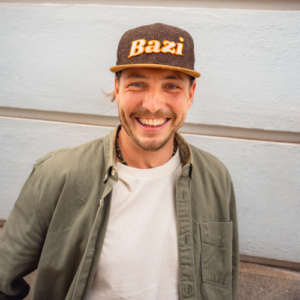 Lächelnder Mann mit grünem Hemd, weißem T-Shirt und dunkelbrauner Cap mit gelbem "Bazi"-Schriftzug steht vor einer hellblauen Wand mit horizontalen Linien.