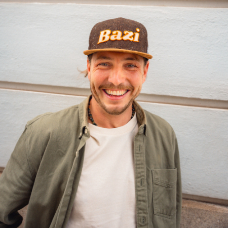 Lächelnder Mann mit grünem Hemd, weißem T-Shirt und dunkelbrauner Cap mit gelbem "Bazi"-Schriftzug steht vor einer hellblauen Wand mit horizontalen Linien.