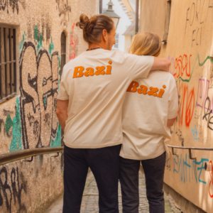 Zwei Personen stehen mit dem Rücken zur Kamera in einer engen Gasse mit Graffiti. Beide tragen beige T-Shirts mit großem orangefarbenem "Bazi"-Schriftzug auf dem Rücken. Die Person links legt den Arm um die andere.