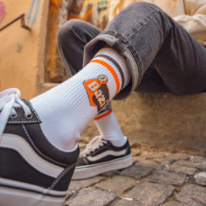 Nahaufnahme eines Sneakers mit hochgezogener weißer Bazi-Socke, auf der das Bazi-Logo in Orange und Schwarz zu sehen ist. Die Person sitzt auf Kopfsteinpflaster in einer schmalen, leicht besprühten Gasse.