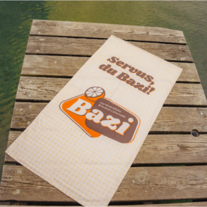 Beiges Bazi-Handtuch mit dem Schriftzug "Servus, du Bazi!" und großem Bazi-Logo liegt ausgebreitet auf einem Holzsteg am Wasser. Der Steg befindet sich über grünlich schimmerndem See.