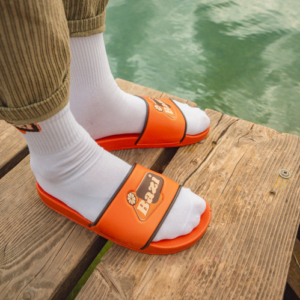 Person steht auf einem hölzernen Steg am Wasser. Sie trägt weiße Sportsocken mit Logo und orangefarbene Bazi-Badelatschen mit auffälligem Logo-Riemen. Dazu eine beige Cordhose.
