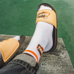 Nahaufnahme von Beinen mit weißen Bazi-Sportsocken und beigefarbenen Bazi-Badelatschen auf einem Holzsteg am Wasser. Die Person trägt schwarze Jeans, im Hintergrund ist die grüne Wasseroberfläche sichtbar.