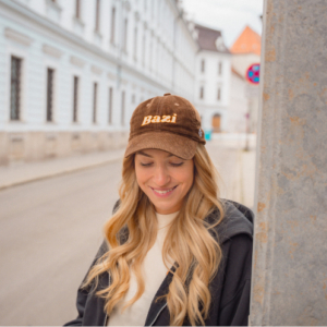 Junge Frau mit langen blonden Haaren steht lächelnd an einer Hauswand in einer Altstadtstraße. Sie trägt eine braune Cord-Cap mit gelbem "Bazi"-Schriftzug.