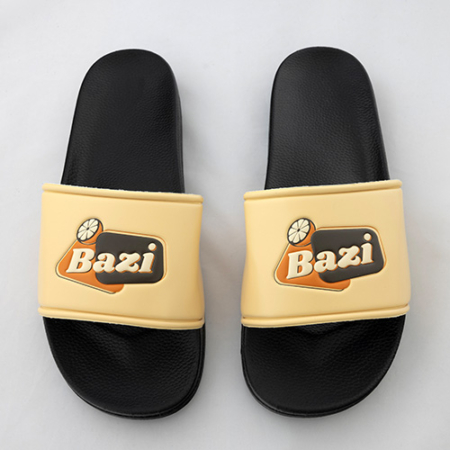 Ein Paar schwarze Badeschlappen mit beigen Riemen. Auf beiden Riemen befindet sich das farbige Bazi-Logo mit Zitronenscheibe. Die Schlappen liegen nebeneinander auf hellem Untergrund.