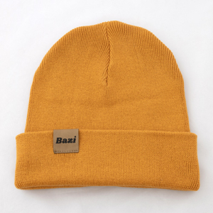 Senfgelbe Strickmütze mit umgeschlagenem Rand. Auf der Vorderseite ist ein kleines, eingenähtes Label mit dem Schriftzug "Bazi" in schwarzer Schrift zu sehen.