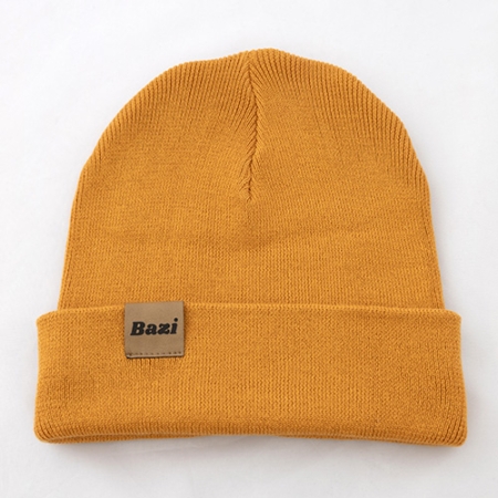 Senfgelbe Strickmütze mit umgeschlagenem Rand. Auf der Vorderseite ist ein kleines, eingenähtes Label mit dem Schriftzug "Bazi" in schwarzer Schrift zu sehen.