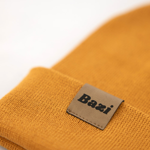 Detailaufnahme einer senfgelben Strickmütze mit umgeschlagenem Rand. Am Saum ist ein kleines braunes Label mit dem schwarzen Schriftzug "Bazi" eingenäht.