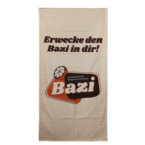 Großes Strandtuch. Cremefarben mit dunkleren Punkten. Großer Schriftzug "Erwecke den Bazi in dir". Darunter befindet sich das Bazi Cola-Mix Logo.