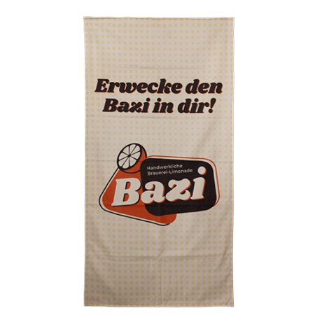 Großes Strandtuch. Cremefarben mit dunkleren Punkten. Großer Schriftzug "Erwecke den Bazi in dir". Darunter befindet sich das Bazi Cola-Mix Logo.