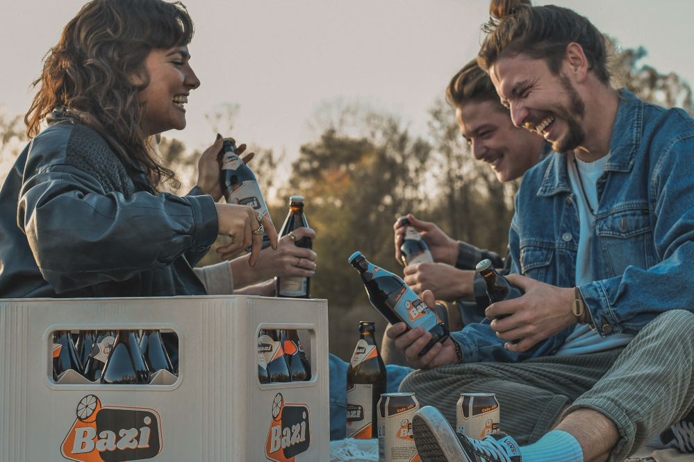 Sitzende Gruppe aus jungen Männern uns Frauen in der frühen Abend Sonne. Vor der Gruppe junger Menschen befindet sich ein Kasten Bazi Cola-Mix mit verschiedenen Sorten.